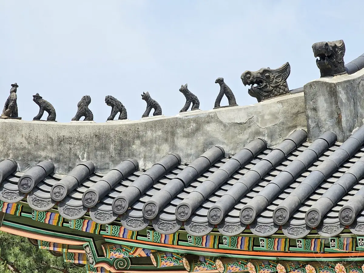 Die Dachlinie des Deoksugung Palace in Seoul, Südkorea, wird von einer Reihe von kunstvollen Tierfiguren und einem imposanten Drachenkopf geschmückt. Die kunstvollen Ziegeldächer und die grauen, wettergegerbten Mauern verleihen der Szene eine historische und ehrwürdige Atmosphäre. Die Szenerie strahlt die traditionelle Ästhetik koreanischer Palastarchitektur aus, die sich eindrucksvoll gegen den hellblauen Himmel abhebt.