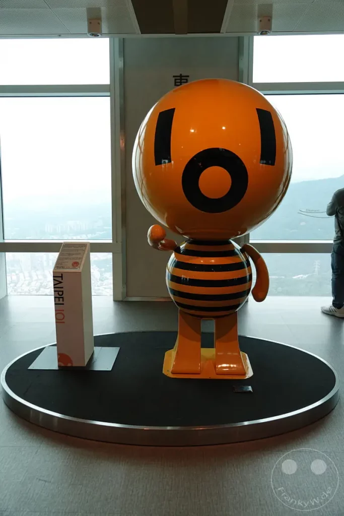 Eine freundliche, orangefarbene Biene oder ein Wesen mit schwarzen Streifen steht auf einer runden, dunklen Ausstellungsfläche vor einem großen Fenster in Taipeh. Hinter dem aufgestellten Maskottchen offenbart sich eine beeindruckende Stadtlandschaft von Taiwan aus der Höhe, die durch die schlichte Gestaltung und das helle Licht eine positive und einladende Atmosphäre vermittelt.