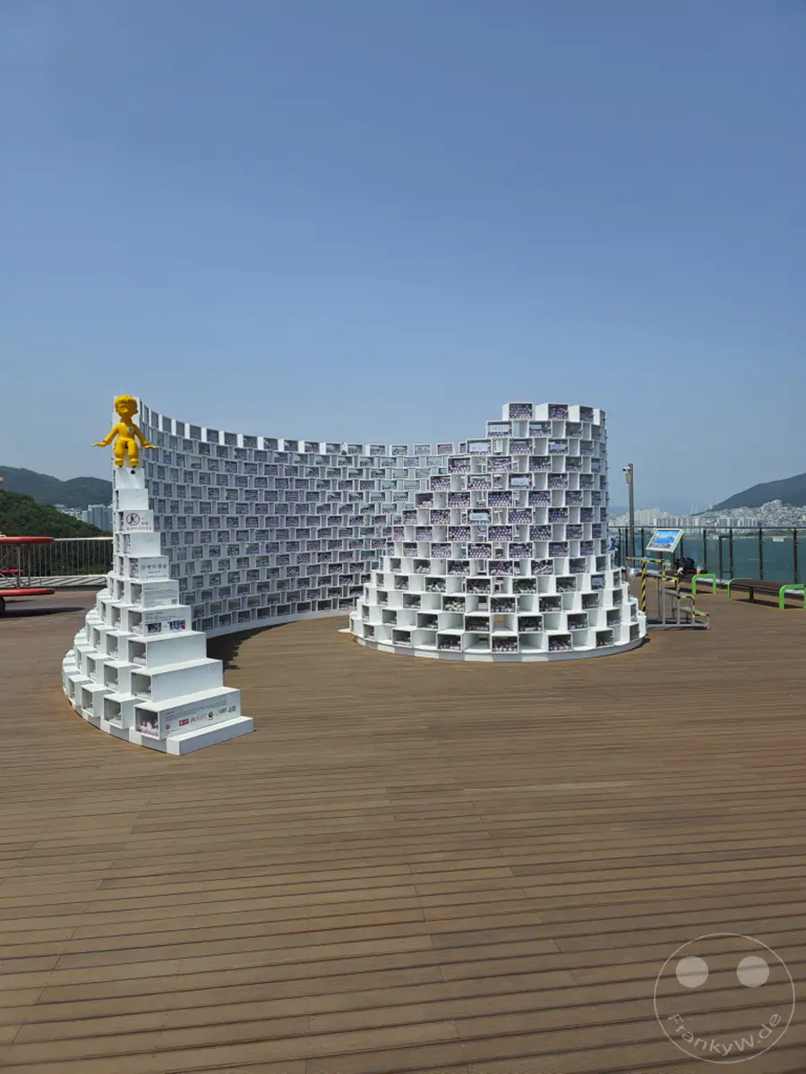 Ein weites Panorama über die Küstenlandschaft Südkoreas in Busan, mit dem Songdo Sky Park im Mittelpunkt. Die geschwungene, modulare Installation aus weißen Boxen und eine kleine gelbe Figur dominieren den hölzernen Boden unter einem klaren blauen Himmel. Die Szene strahlt eine moderne, ruhige Atmosphäre aus, die zum Verweilen einlädt.