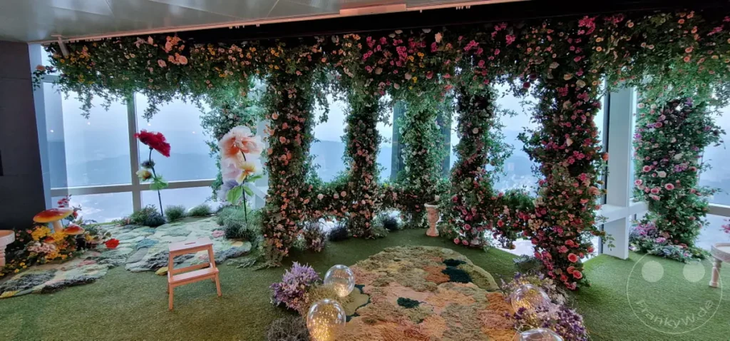 Eine üppige Blumendekoration umrahmt die Fensterfront mit Blick auf Taipeh, Taiwan, und die Skyline von Taipei 101. Ein verträumter Garten mit künstlichen Pilzen und Moos-Elementen schafft eine märchenhafte Atmosphäre. Die Szenerie eignet sich perfekt für besondere Anlässe und Events mit beeindruckendem Ausblick.
