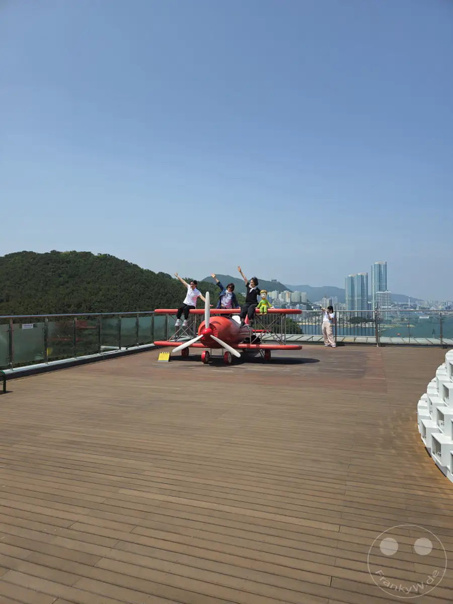 Eine Gruppe von vier jungen Frauen posiert fröhlich auf und neben einem roten Flugzeugspielgerät auf der Aussichtsplattform des Songdo Sky Park in Busan, Südkorea. Im Hintergrund erstreckt sich die Stadt mit ihren Hochhäusern und dem weiten Meer, eingerahmt von grünen Hügeln. Das sonnige Wetter und die entspannte Atmosphäre laden zum Verweilen und Genießen des Panoramablicks ein.