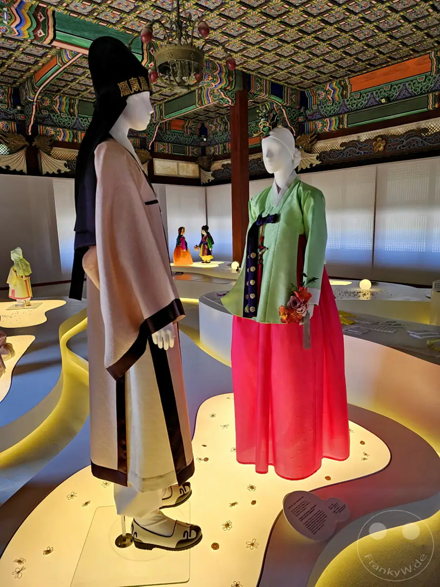 Im Deoksugung Palace in Seoul werden traditionelle koreanische Hanboks auf kunstvollen Podesten präsentiert. Die farbenprächtigen Gewänder strahlen eine Aura der Geschichte und kulturellen Bedeutung aus, die Besucher in die Vergangenheit eintauchen lässt. Eine detailreiche Deckenbemalung rundet die immersive Ausstellung ab.