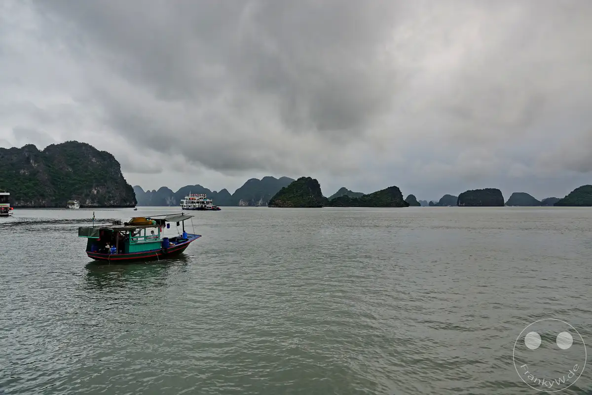 Eine düstere Wolkendecke liegt über der berühmten Ha Long Bucht in Vietnam, während sich die ruhigen Gewässer unter ihr spiegeln. Mehrere Boote, darunter ein kleineres Fischerboot im Vordergrund und ein größeres Ausflugsboot weiter hinten, sind auf dem Wasser unterwegs. Die Szene vermittelt eine melancholische und ruhige Atmosphäre, die durch die dramatische Wolkenlandschaft und die Silhouette der Karstinseln noch verstärkt wird.