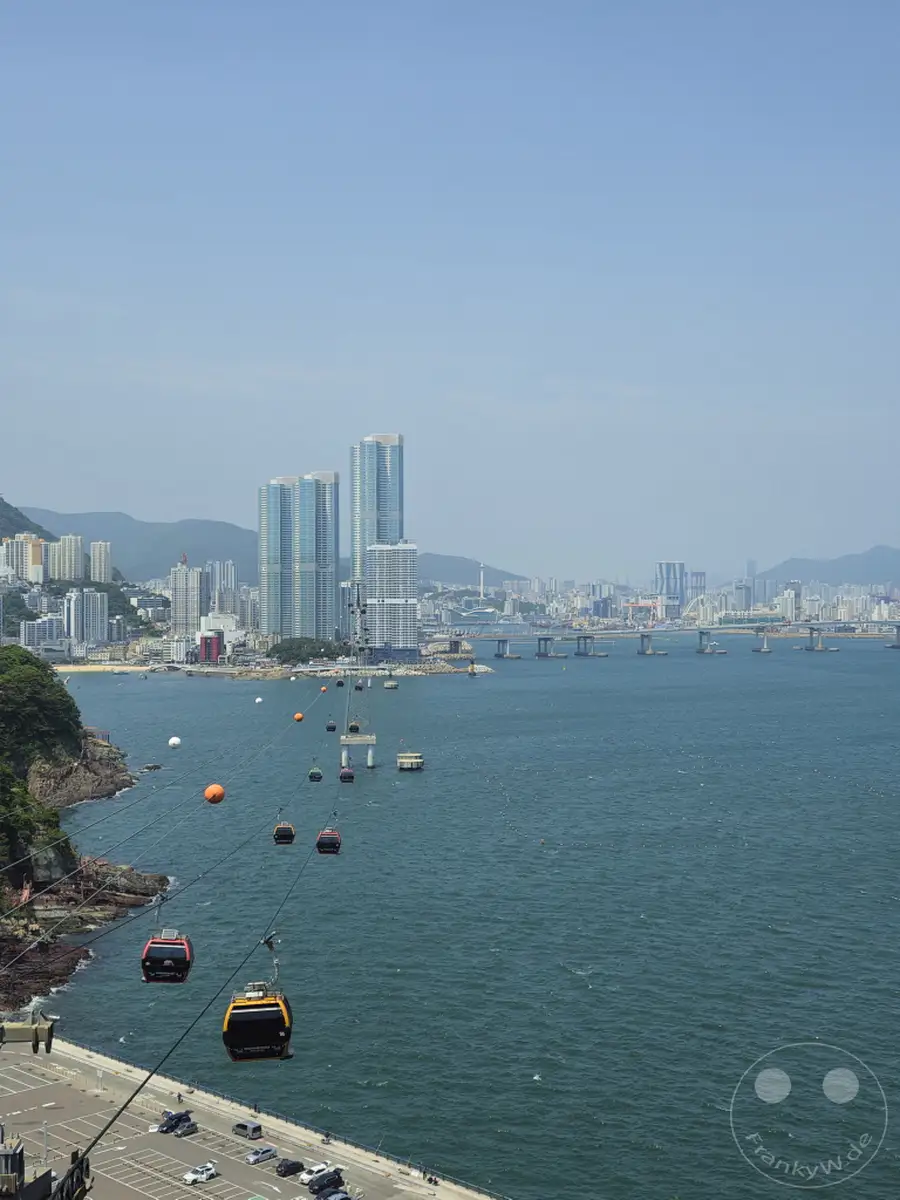 In Busan, Südkorea, gleiten die Gondeln des Songdo Sky Parks über das tiefblaue Meer, mit Blick auf die beeindruckende Skyline der Stadt und die weitläufige Brücke. Die Seilbahn bietet eine spektakuläre Perspektive auf die Küstenlandschaft und das geschäftige Treiben unterhalb. Sonnenschein und die klare Luft erzeugen eine entspannte und maritime Atmosphäre.