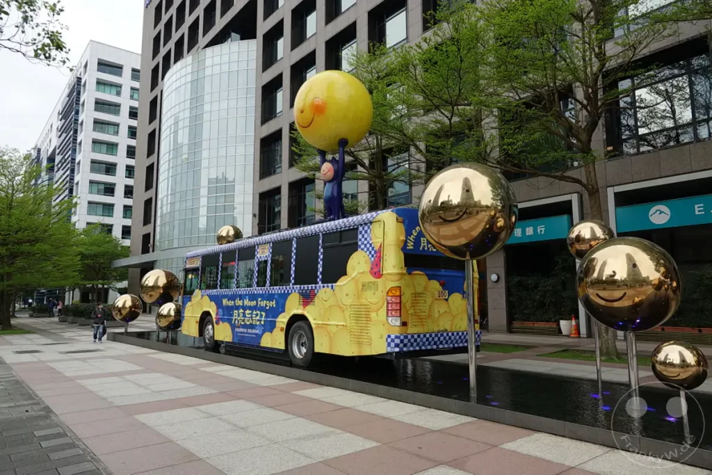 In Taipeh, Taiwan, steht ein thematischer Bus, der zur Kunstinstallation "When the Moon Forgot" gehört. Mehrere spiegelnde Kugelskulpturen mit Smiley-Gesichtern sind in der Nähe aufgestellt und reflektieren die moderne Architektur der Stadt. Die Szene wirkt lebendig und verspielt, inmitten eines urbanen Umfelds.
