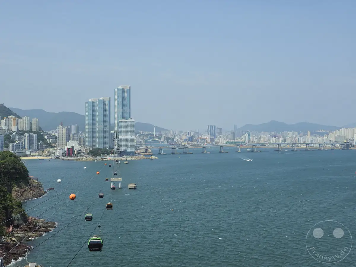 Eine weite Panoramaaufnahme des Hafens von Busan, Südkorea, zeigt die Songdo-Seilbahn, die über das glitzernde Meer schwebt und zwei Ufer verbindet. Im Hintergrund erhebt sich die beeindruckende Skyline der Stadt mit zahlreichen Hochhäusern, während eine massive Brücke die Küste überspannt. Die Szene strahlt eine lebendige Atmosphäre der modernen Metropole und der maritime Schönheit aus.