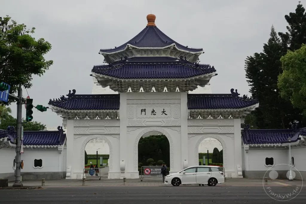 Die imposante Architektur der Chiang Kai-shek Memorial Hall in Taipeh, Taiwan, prägt das Bild unter einem bewölkten Himmel. Weiße Gebäude mit blauen Dächern und chinesischen Schriftzeichen schaffen eine ehrfürchtige Atmosphäre. Eine weiße Limousine parkt vor dem Eingang, während vereinzelte Besucher den Ort erkunden.