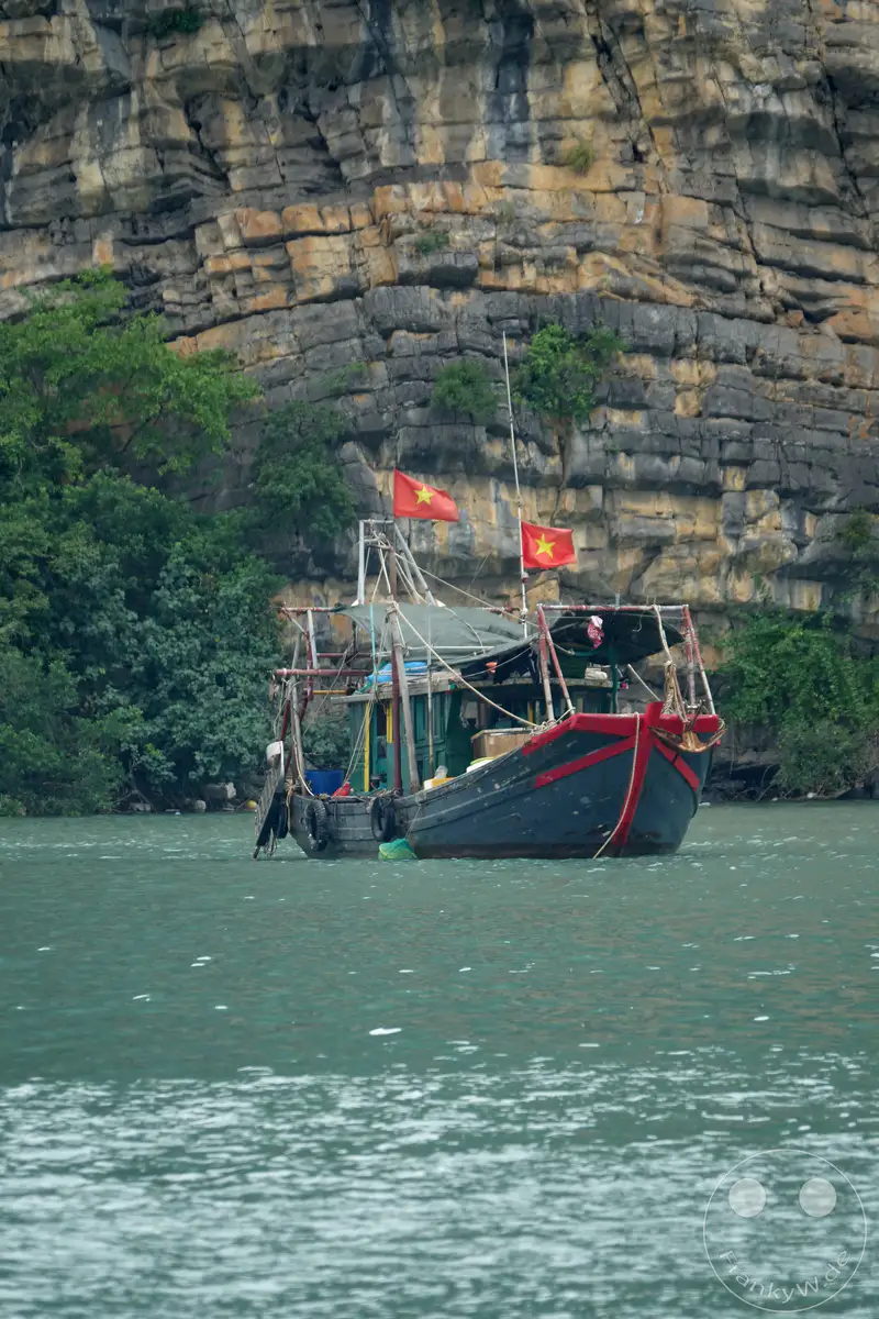 Ein traditionelles vietnamesisches Fischerboot ankert ruhig im smaragdgrünen Wasser der weltberühmten Halong-Bucht in Vietnam. Im Hintergrund ragen majestätisch zerklüftete Karstfelsen aus dem Meer auf, die von üppiger grüner Vegetation bedeckt sind. Die Szene strahlt eine friedliche und naturbelassene Atmosphäre aus, die typisch für diese ikonische Landschaft ist.