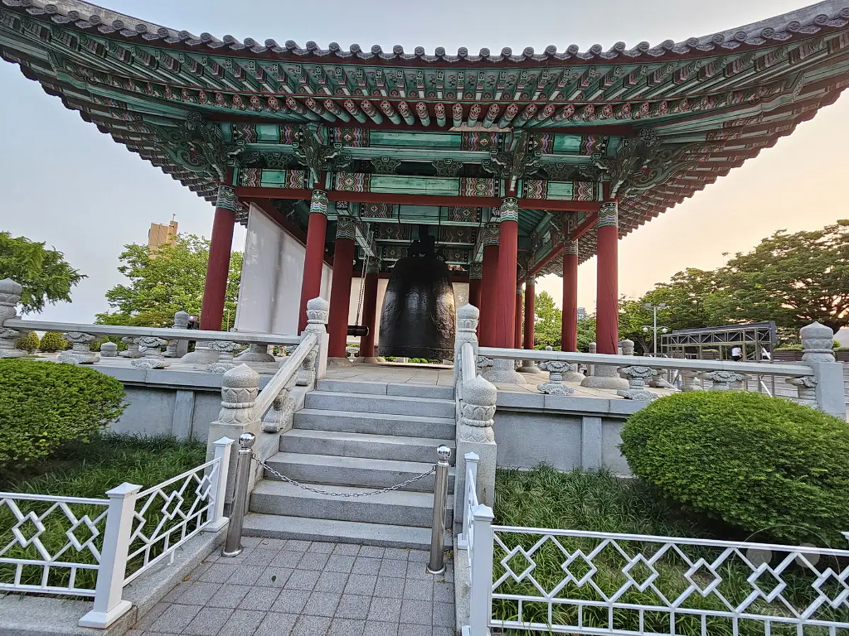 Ein imposanter Glockenturm im traditionell koreanischen Stil thront in Südkoreas Busan auf dem Yongdusan Park. Die kunstvoll gestaltete Dachkonstruktion und die massive Glocke im Zentrum des Pavillons strahlen Ruhe und kulturelle Bedeutung aus. Umgeben von gepflegten Grünflächen und einer stoneinen Balustrade, vermittelt die Szene eine friedliche und erhabene Atmosphäre.