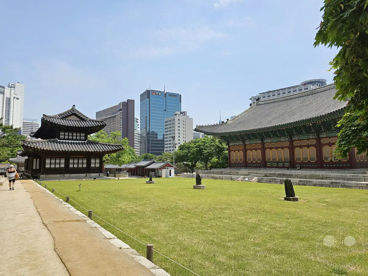 Ein Blick über eine gepflegte Rasenfläche auf die historische Architektur des Deoksugung Palastes in Seoul, Südkorea. Im Hintergrund überragen moderne Hochhäuser die traditionellen Gebäude und bilden einen faszinierenden Kontrast. Die Szene strahlt eine friedliche Atmosphäre unter einem strahlend blauen Himmel aus.