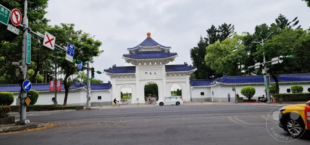 Die beeindruckende Toranlage der Chiang Kai-shek Memorial Hall in Taipeh, Taiwan, erhebt sich mit ihrer traditionellen blauen Dachziegelarchitektur. Auf der belebten Kreuzung vor dem Tor bewegen sich Fahrzeuge und Radfahrer im städtischen Treiben unter einem bedeckten Himmel. Die Szene strahlt eine Mischung aus historischer Bedeutung und alltäglichem Leben in der Hauptstadt Taiwans aus.
