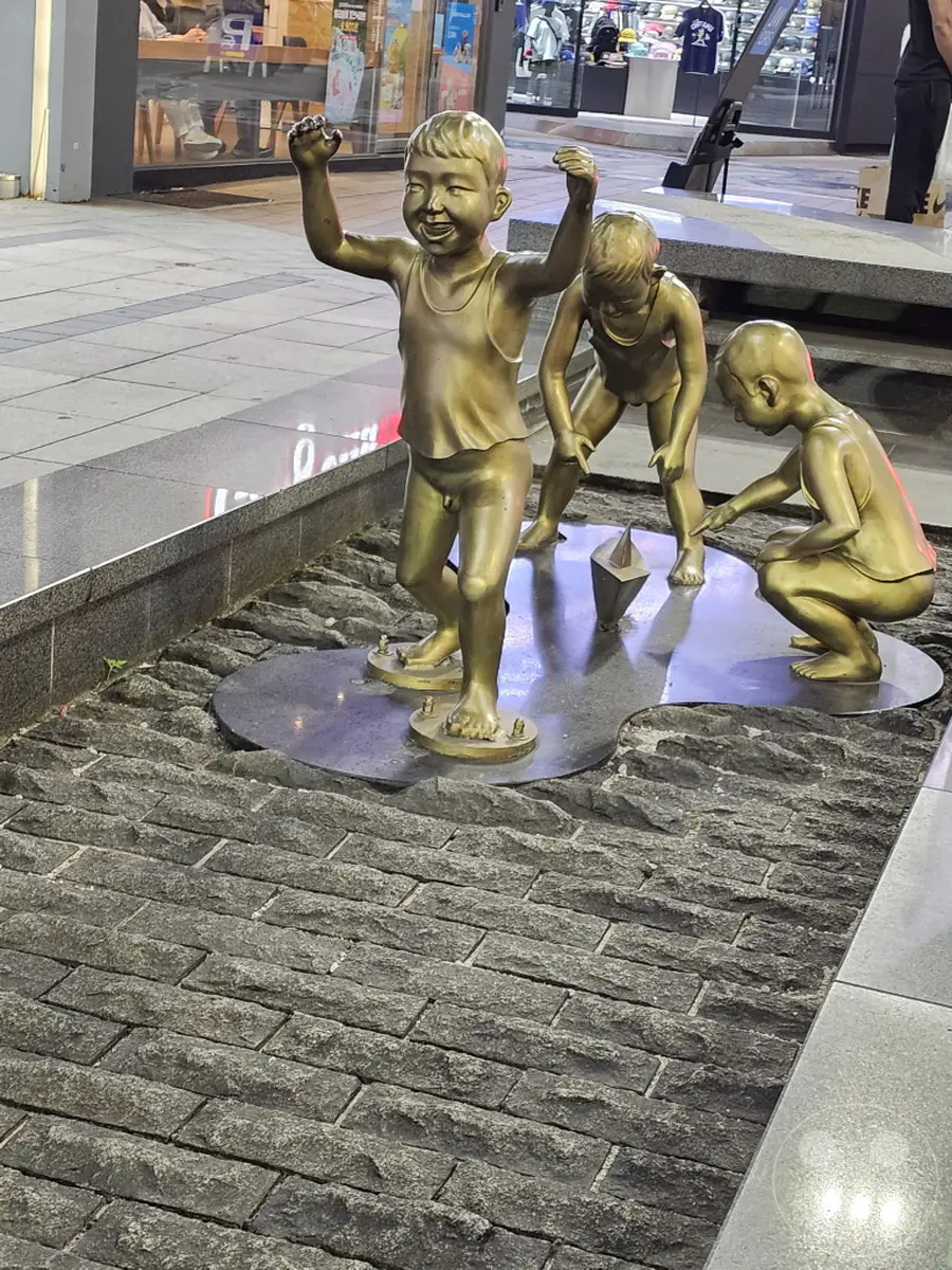Drei bronzene Kinderskulpturen spielen in Südkorea, Busan, auf der Gwangbok-ro street mit Papierbooten. Die lebensechten Figuren vermitteln eine fröhliche und spielerische Stimmung, während sie sich auf einem künstlichen Gewässer tummeln. Der umliegende gepflasterte Bereich deutet auf einen öffentlichen Platz oder eine Einkaufsstraße hin.