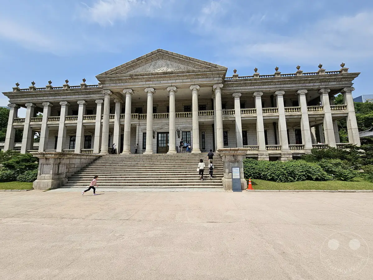 Der Deoksugung Palace in Seoul, Südkorea, präsentiert sich als beeindruckendes Beispiel historischer Architektur mit seinen imposanten Säulen und weitläufigen Treppen. Die Szene strahlt eine Mischung aus majestätischer Ruhe und lebendiger Atmosphäre aus, während Besucher die Anlage erkunden. Ein junges Mädchen rennt fröhlich über den Vorplatz und verleiht dem historischen Ort eine moderne, spielerische Note.