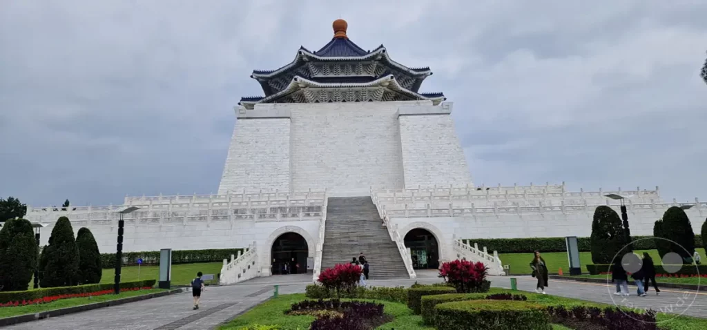 Das Chiang Kai-shek Memorial Hall in Taipeh, Taiwan, ist ein imposantes Bauwerk mit traditioneller chinesischer Architektur und einem umliegenden Park. Die weiße Marmorfassade und die blauen Dächer verleihen dem Monument eine feierliche Ausstrahlung, während die gepflegten Grünflächen eine friedliche Atmosphäre schaffen. Menschen bewegen sich auf dem weitläufigen Gelände und erkunden die Gedenkstätte an einem bewölkten Tag.