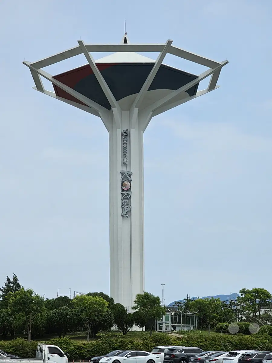 Ein imposanter weißer Turm, der mit den Worten „Welcome to KOREA“ und dem Emblem Südkoreas verziert ist, dominiert den Himmel. Darunter erstreckt sich ein grüner Hang mit Büschen und Bäumen, während am unteren Bildrand eine Reihe von Autos parkt. Die Szenerie am Gimhae International Airport in Busan vermittelt ein Gefühl der Ankunft und des Willkommens.