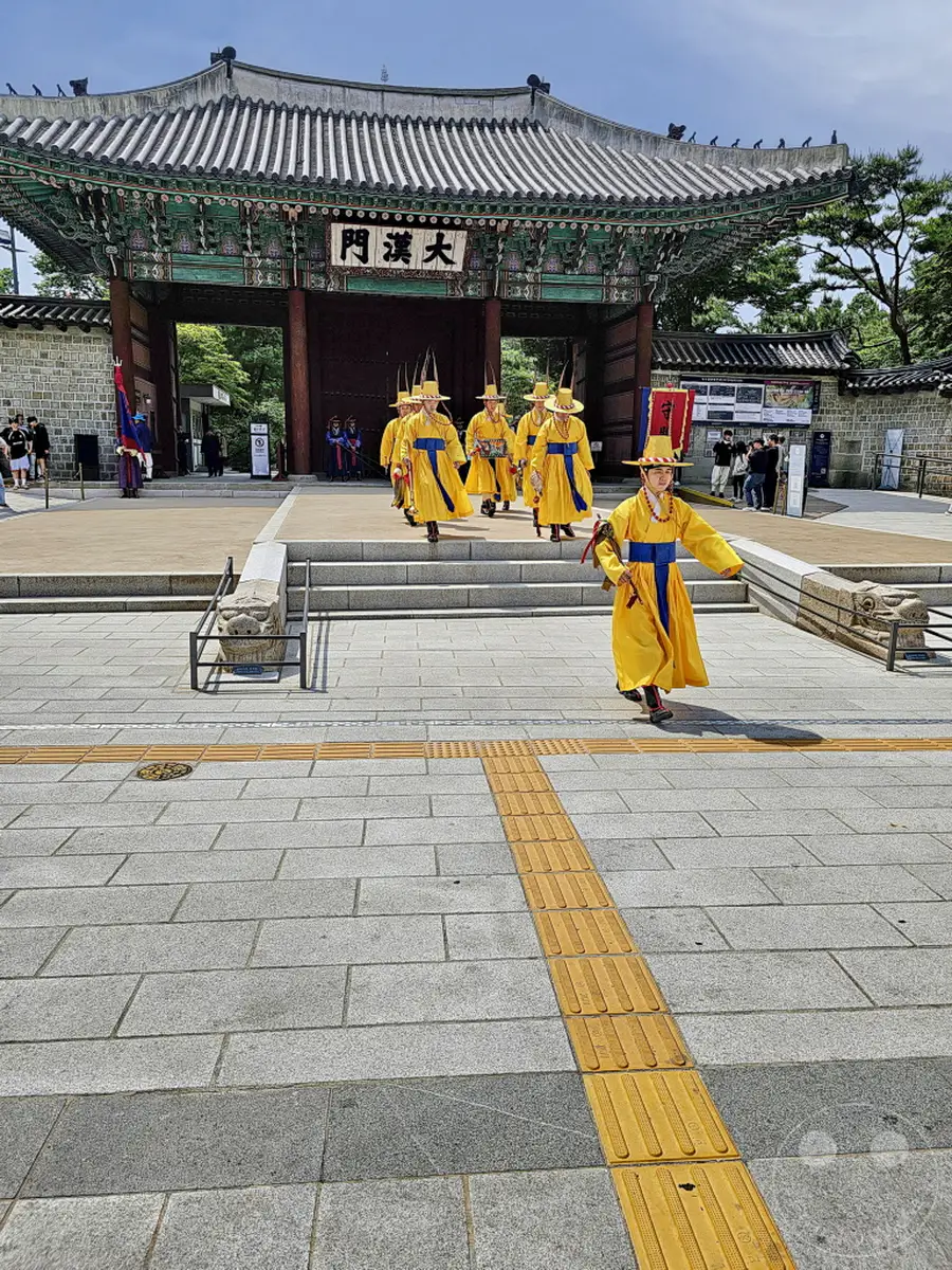 In Seoul, Südkorea, findet am Deoksugung Palace eine traditionelle Wachablösungszeremonie statt. Wachen in leuchtend gelben Gewändern und traditionellen Hüten marschieren den Eingangsbereich entlang, der von historischer Architektur geprägt ist. Die Szene vermittelt ein Gefühl von kulturellem Erbe und feierlicher Prozession vor dem beeindruckenden Palasttor.