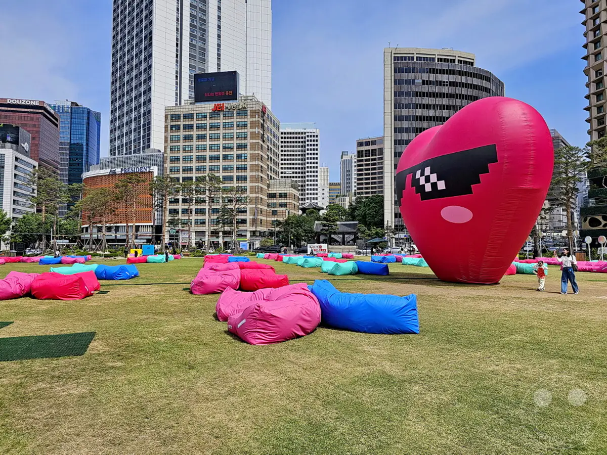 Ein riesiger, aufblasbarer Herzballon mit pixeliger Sonnenbrille steht auf einer Grünfläche in Seoul, Südkorea. Über den Rasen sind zahlreiche bunte Sitzsäcke verteilt, die zum Entspannen einladen. Im Hintergrund erheben sich imposante Hochhäuser, die einen Kontrast zur entspannten Atmosphäre im Park bilden.