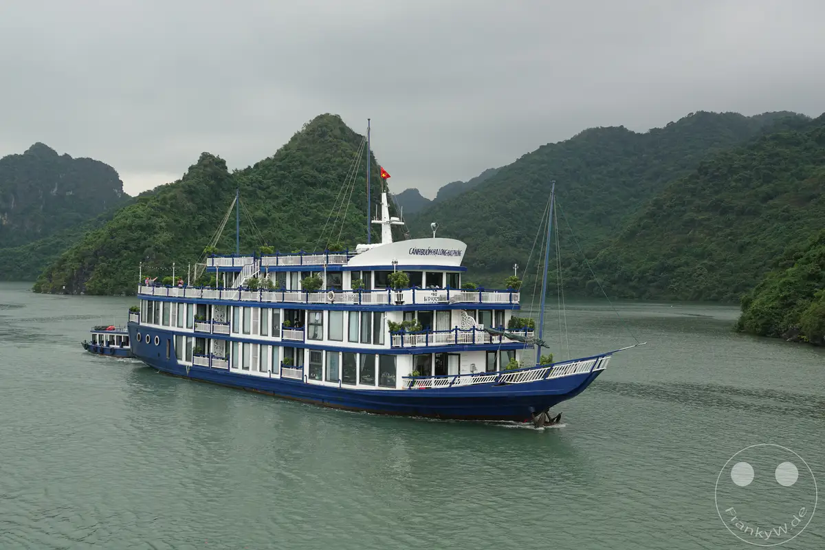 Eine blau-weiße Dschunke gleitet durch die ruhigen Gewässer der Ha Long Bucht in Vietnam. Üppig grüne Karstfelsen ragen majestätisch aus dem Wasser und bilden eine atemberaubende Kulisse für die maritime Szenerie. Die Wolken am Himmel sorgen für eine gedämpfte, friedliche Atmosphäre, die zum Entspannen einlädt.
