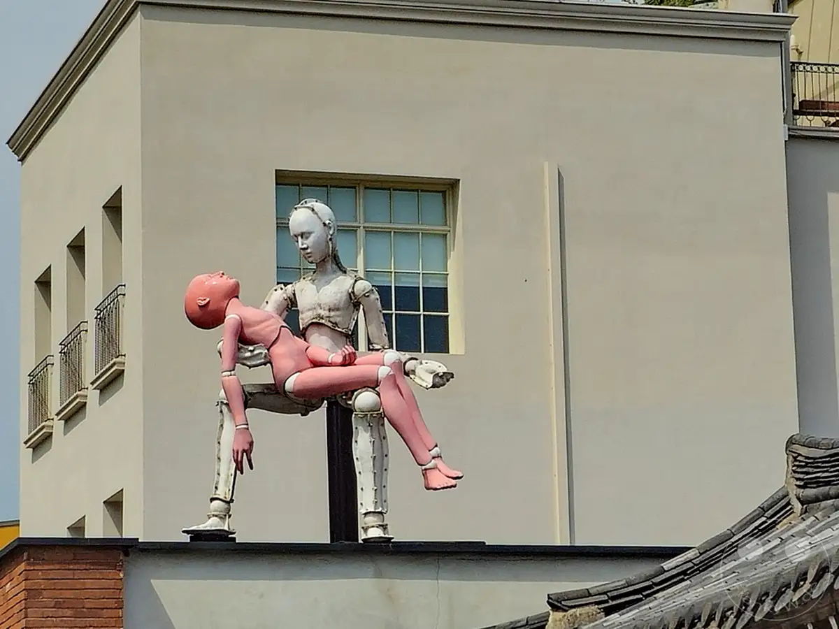 Eine Kunstinstallation mit zwei Roboterfiguren ist vor der Hakgojae Gallery in Seoul, Südkorea, zu sehen. Eine menschenähnliche Skulptur hält eine kleinere, rosa dargestellte Figur in ihren Armen. Die Szenerie wirkt nachdenklich und stellt möglicherweise eine Verbindung zwischen Technologie und Menschlichkeit dar.