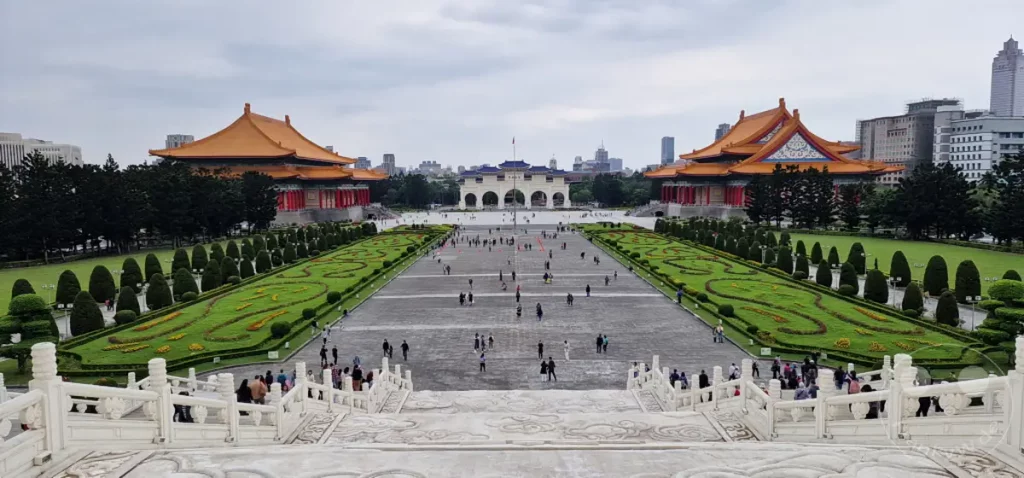 Der weite Platz vor der Chiang Kai-shek Memorial Hall in Taipeh, Taiwan, erstreckt sich unter einem bewölkten Himmel. Zu beiden Seiten flankieren das Nationaltheater und der Nationalkonzertsaal die symmetrisch angelegten Grünflächen und den zentralen Durchgang. Menschen spazieren über den Platz und genießen die beeindruckende Architektur und die parkähnliche Anlage.