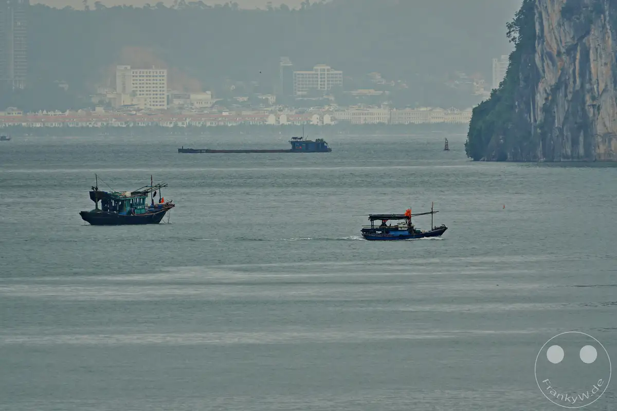 Zwei traditionelle Boote, eine Dschunke und ein Fischerboot, gleiten über das ruhige Wasser der Ha Long Bucht in Vietnam. Im Hintergrund erhebt sich eine imposante Karstinsel vor einer nebligen Küstenstadt. Die Szenerie strahlt eine friedliche und majestätische Atmosphäre aus, die die natürliche Schönheit der Region hervorhebt.