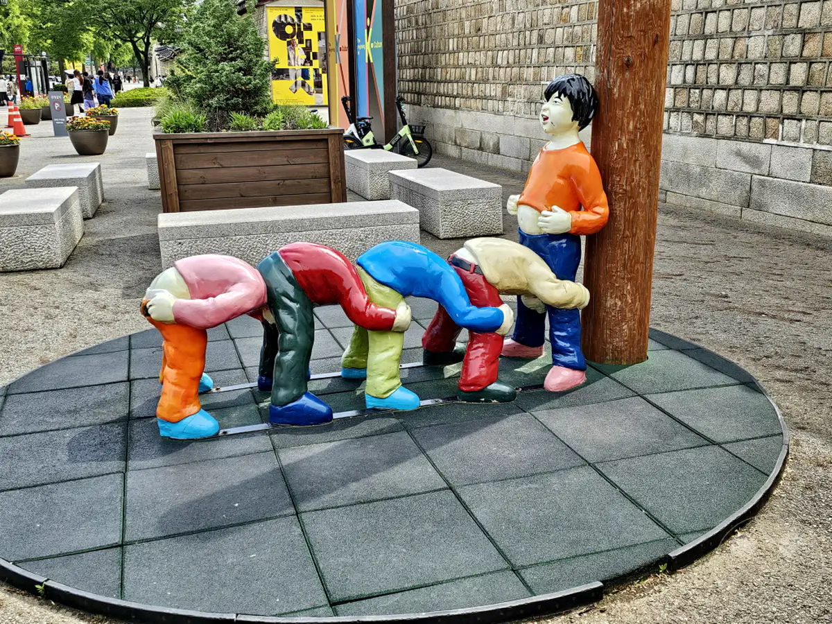 In Seoul, Südkorea, ist eine humorvolle Skulpturengruppe vor dem Gyeongbokgung Palace zu sehen. Vier Kinderfiguren sind dargestellt, wie sie spielerisch den Kopf zwischen die Beine stecken, während eine fünfte Figur mit einem breiten Lächeln daneben steht. Die Szene vermittelt eine ausgelassene und fröhliche Atmosphäre auf einem öffentlichen Platz.