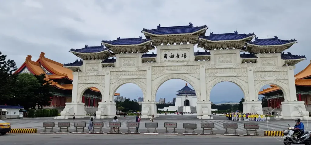 Der imposante Liberty Square Arch in Taipeh, Taiwan, markiert den Eingang zum Nationalen Freiheitsplatz. Die traditionelle chinesische Architektur mit ihren blauen Dächern und weißen Steinreliefs dominiert die Szenerie unter einem bedeckten Himmel. Menschen sind auf dem Platz unterwegs und schaffen eine belebte, aber respektvolle Atmosphäre vor dem beeindruckenden Bauwerk.