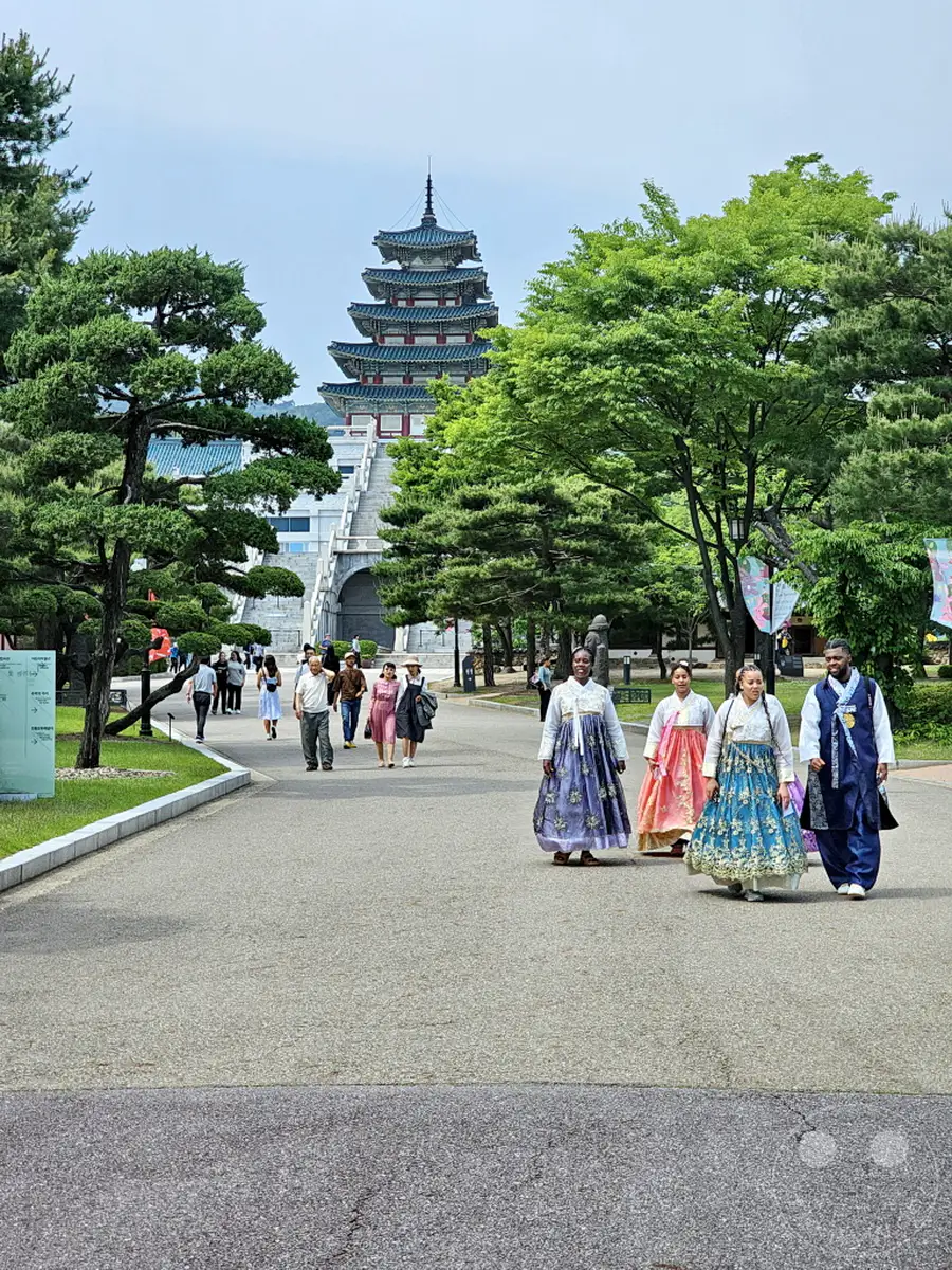 Eine Gruppe von Personen in traditioneller koreanischer Kleidung (Hanbok) spaziert auf einem Weg zum Gyeongbokgung Palast in Seoul, Südkorea. Im Hintergrund erhebt sich die beeindruckende Architektur des Palastes, umgeben von üppigem Grün. Die Szene vermittelt ein Gefühl von kulturellem Erbe und friedlicher Erkundung.