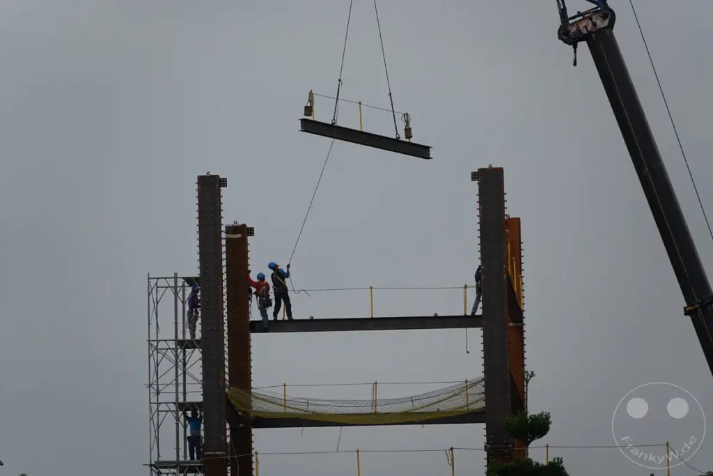 In Taipeh, Taiwan, schreiten die Bauarbeiten eines neuen Stahlkonstrukts voran, während Arbeiter mit einem Kran einen schweren Stahlträger positionieren. Ein Gerüst und Sicherheitsnetze sind sichtbar und unterstreichen die sorgfältige Planung und Ausführung des Bauprojekts unter einem bewölkten Himmel. Die Szene vermittelt ein Gefühl von Fortschritt und menschlicher Anstrengung im industriellen Sektor.
