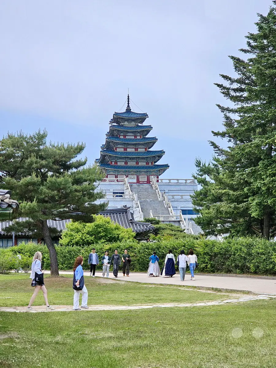 Eine Gruppe von Menschen spaziert auf einem Weg durch den Gyeongbokgung Palast in Seoul, Südkorea. Im Hintergrund erhebt sich die beeindruckende, mehrstöckige Pagode vor einem bewölkten Himmel. Üppiges Grün und alte Bäume säumen den Weg und verleihen der Szene eine friedliche Atmosphäre.