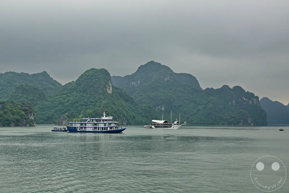 Die Ha Long Bucht in Vietnam ist berühmt für ihre beeindruckenden Karstfelsen, die aus dem smaragdgrünen Wasser ragen. Mehrere Schiffe, darunter eine traditionelle Dschunke, kreuzen auf der ruhigen See vor der malerischen Kulisse. Eine mystische, leicht diesige Atmosphäre liegt über der Landschaft.