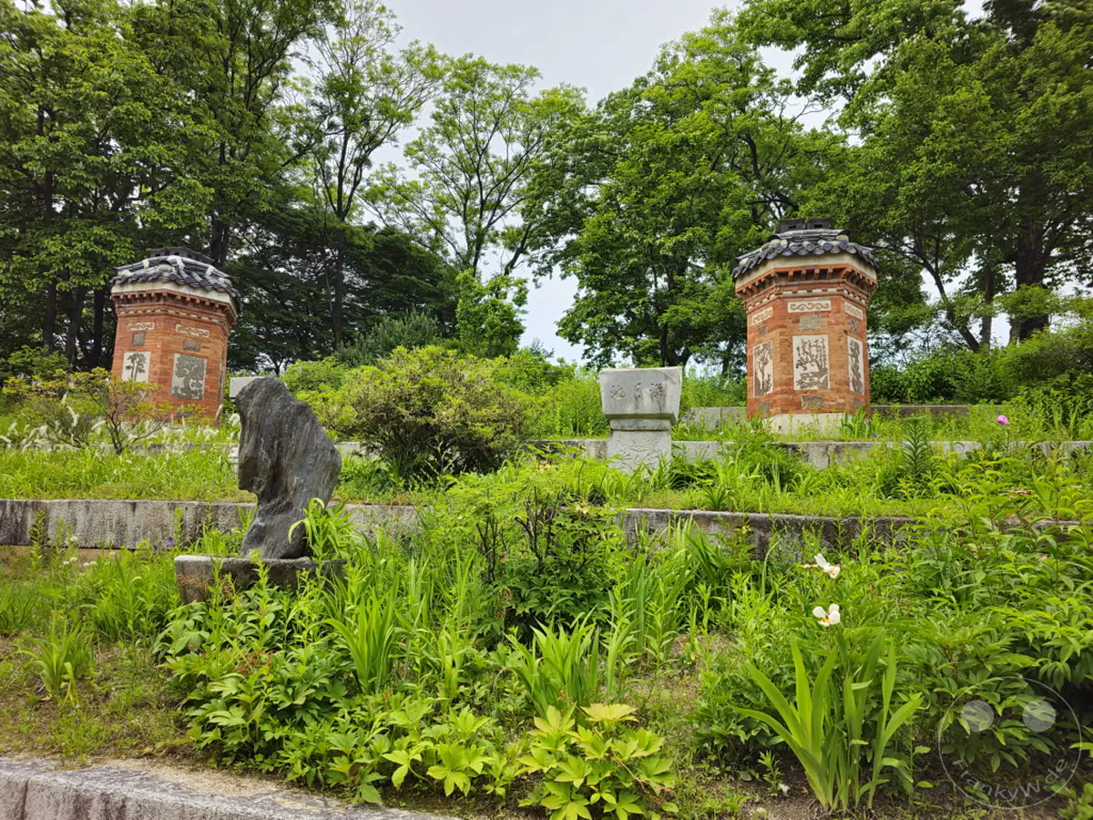 Zwei achteckige Ziegelgebäude mit traditionellen Dächern stehen auf einer ansteigenden, begrünten Fläche im Gyeongbokgung Palast in Seoul, Südkorea. Dazwischen und davor hebt sich eine steinerne Skulptur ab, umgeben von üppigem Grün und Blumen. Die Szene vermittelt eine ruhige und historisch bedeutsame Atmosphäre inmitten der Natur.