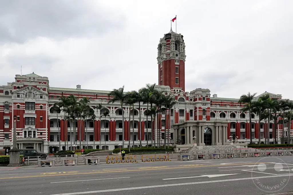 Das imposante Gebäude des Presidential Office Building in Taipeh, Taiwan, präsentiert sich in seiner roten und weißen Architektur unter einem bedeckten Himmel. Vor dem Gebäude stehen hohe Palmen, die dem Anblick eine tropische Note verleihen. Die Szene wirkt würdevoll und zeugt von der historischen Bedeutung des Ortes.