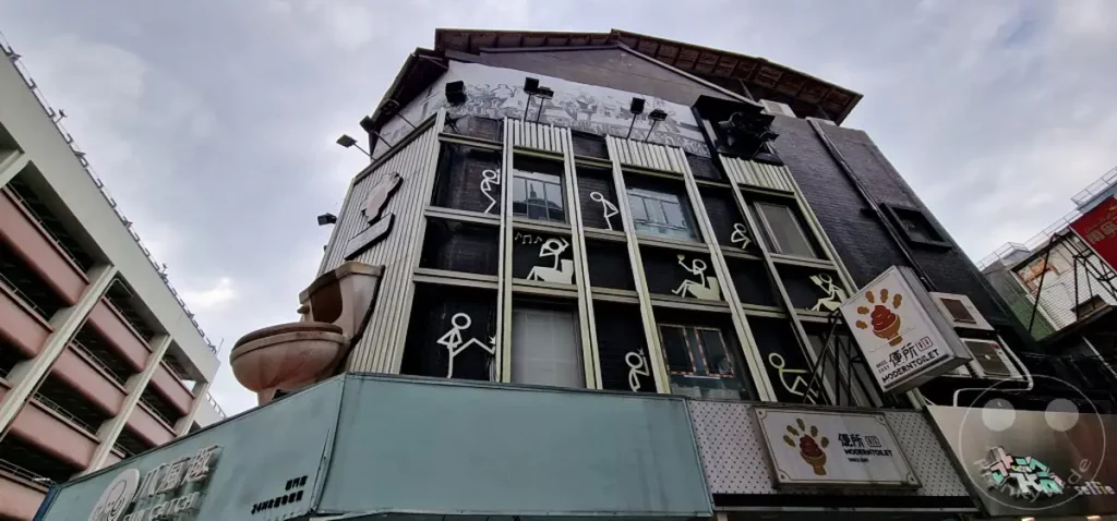 Ein skurriles Gebäude in Taipeh, Taiwan, präsentiert eine ungewöhnliche Fassadengestaltung mit einer prominenten Toilettenskulptur und stickfigurartigen Dekorationen. Die Architektur spiegelt einen humorvollen und verspielten Ansatz wider, der Neugier weckt und ein einzigartiges visuelles Erlebnis bietet. Überwiegend grauer Himmel bildet einen Kontrast zur auffälligen und fantasievollen Gestaltung des "ModernToilet" Restaurants.
