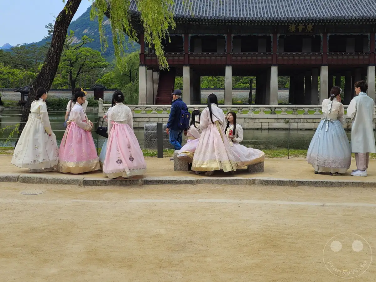 Mehrere Personen in traditionellen koreanischen Hanbok-Kleidern stehen und sitzen in der Nähe eines Teiches vor dem Gyeongbokgung Palast in Seoul. Im Hintergrund ist ein Teil des Palastgebäudes mit seinen charakteristischen dunklen Holzsäulen und dem Dach zu sehen. Die Atmosphäre ist ruhig und spiegelt das Interesse der Besucher an der koreanischen Kultur und Geschichte wider.