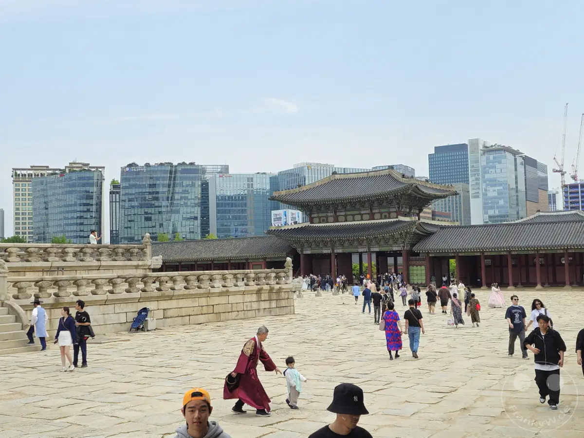 Der Gyeongbokgung Palast in Seoul, Südkorea, präsentiert eine faszinierende Mischung aus historischer Architektur und modernen Hochhäusern im Hintergrund. Touristen schlendern über den weitläufigen Platz und bewundern die prachtvollen Gebäude und die umliegende Landschaft. Die Szene strahlt eine friedliche Atmosphäre aus, die die reiche Geschichte und die dynamische Gegenwart der Stadt widerspiegelt.