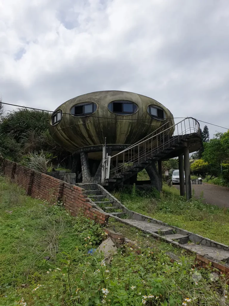 Ein verlassenes UFO-Haus im Wanli District von Taiwan, auch bekannt als Futuro Village. Die einzigartige Architektur steht einsam inmitten üppiger Vegetation und verwitterter Ziegelmauern, was eine melancholische und nostalgische Atmosphäre erzeugt. Überreste von Treppen und Wegen deuten auf frühere Besucher hin, während der trübe Himmel die verlassene Stimmung unterstreicht.