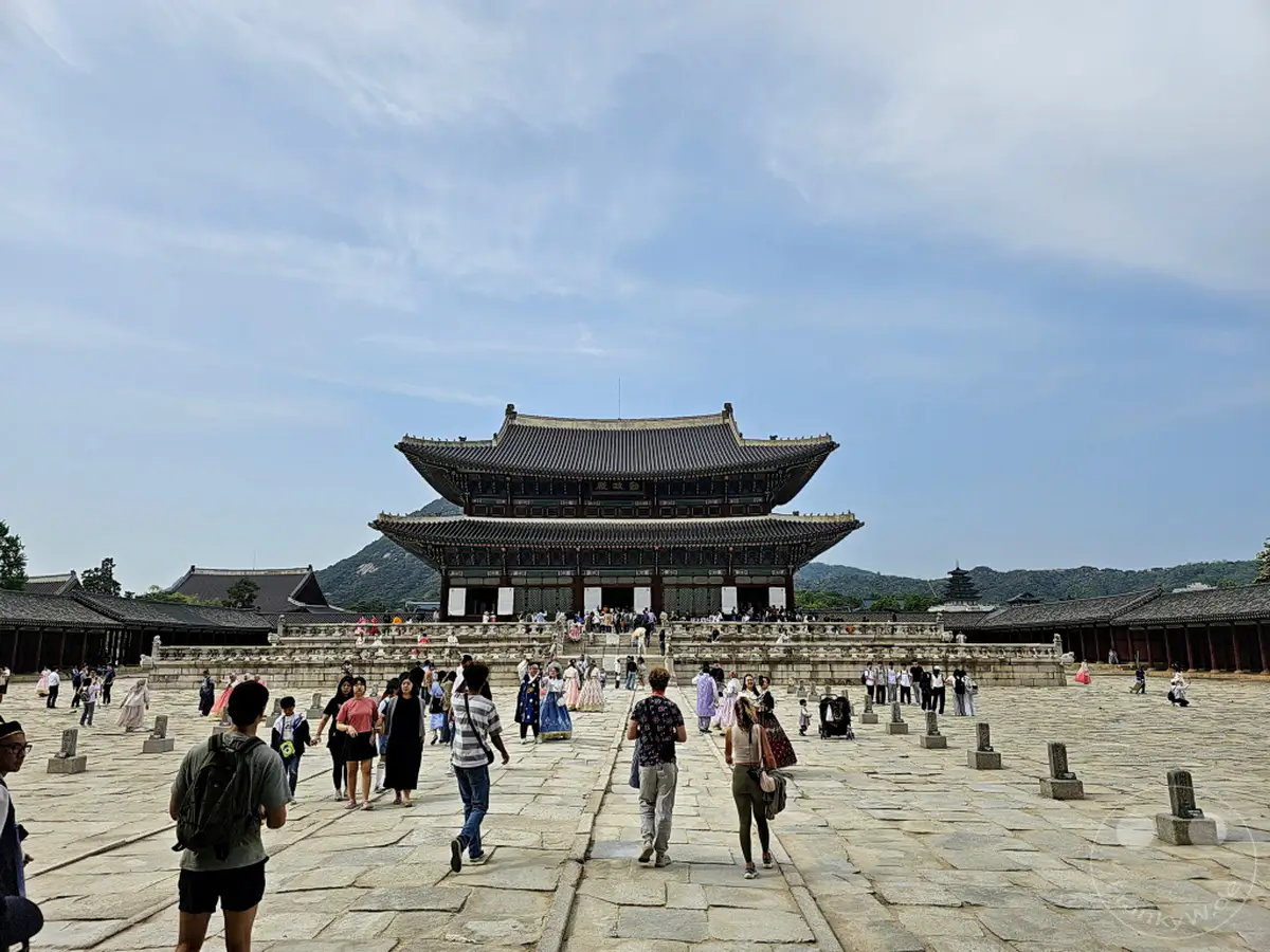 Besucher tummeln sich im weitläufigen Hof des Gyeongbokgung Palastes in Seoul, Südkorea. Die traditionelle koreanische Architektur des Hauptgebäudes und die umliegenden Strukturen bilden eine beeindruckende Kulisse für das geschäftige Treiben. Eine heitere Atmosphäre herrscht unter dem weiten Himmel, während die Menschen die historische Stätte erkunden.