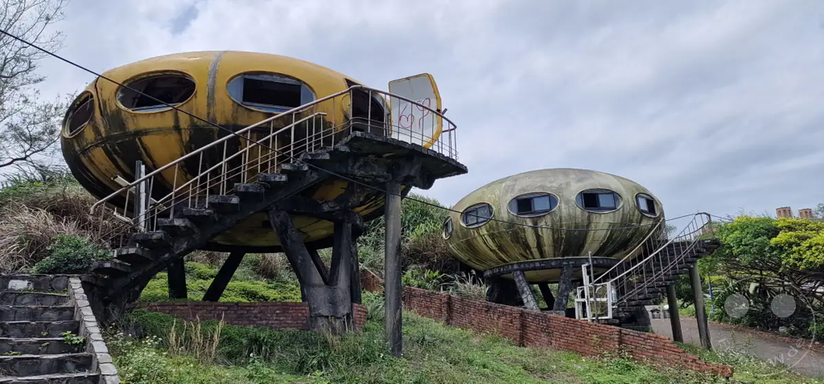 Zwei verlassene Futuro-Häuser, bekannt als das "UFO Village", stehen in Wanli, Taiwan. Die ungewöhnliche Architektur und der verwitterte Zustand der Gebäude erzeugen eine melancholische und surreale Atmosphäre. Umgeben von üppiger Vegetation und unter einem bewölkten Himmel, spiegeln die Strukturen eine vergessene Vision der Zukunft wider.