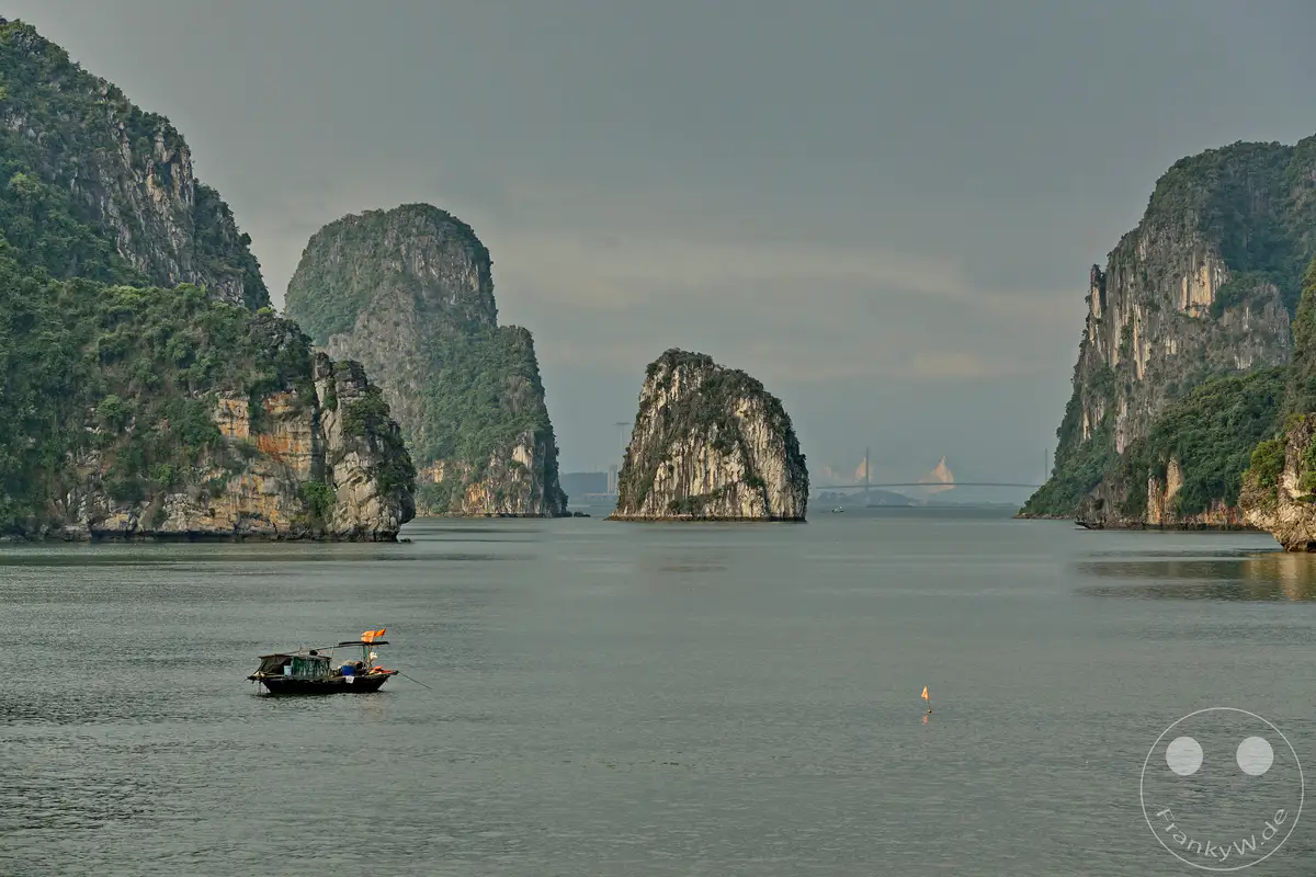 In der Ha Long Bucht Vietnams ragen majestätische Kalksteinfelsen aus dem ruhigen Meer empor, die von üppiger Vegetation bedeckt sind. Eine kleine Holzfähre mit einer orangefarbenen Flagge treibt auf dem trüben Wasser, während im Hintergrund eine weit entfernte Brücke und städtische Silhouetten die Szenerie prägen. Die gedämpfte Beleuchtung und die graue, bewölkte Himmel verleihen der beeindruckenden Landschaft eine mystische und nachdenkliche Atmosphäre.