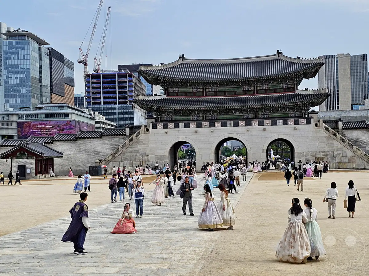Besucher in traditionellen Hanbok-Kleidern erkunden den Gyeongbokgung Palast in Seoul, Südkorea. Die historische Architektur des Palastes bildet einen eindrucksvollen Kontrast zu den modernen Gebäuden im Hintergrund. Eine lebhafte Atmosphäre herrscht auf dem weitläufigen Platz, während Touristen die Sehenswürdigkeit genießen und fotografieren.