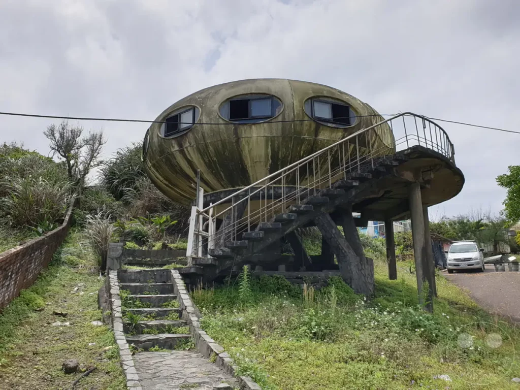 Das verlassene UFO Village in Wanli, Taiwan, beherbergt eine Reihe von surrealen Futuro-Häusern, die auf einem Hügel mit Blick auf die Küste stehen. Die architektonischen Kuriositäten sind von üppigem Grün und einer leicht melancholischen Atmosphäre umgeben, die von ihrem fortschreitenden Verfall zeugt. Eine steile Steintreppe führt zu den ungewöhnlichen, kuppelförmigen Strukturen, während im Hintergrund eine Straße und ein geparktes Auto zu sehen sind.