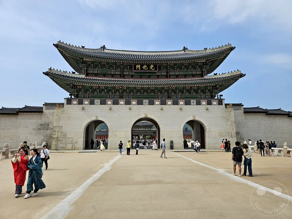 Das Gyeongbokgung Palace in Seoul, Südkorea, präsentiert sich mit seiner prächtigen Haupttorarchitektur unter einem blauen Himmel. Menschen in traditioneller Kleidung und moderne Touristen bewegen sich auf dem weitläufigen Platz vor dem beeindruckenden Bauwerk, das eine Mischung aus historischem Erbe und lebendigem Treiben widerspiegelt.