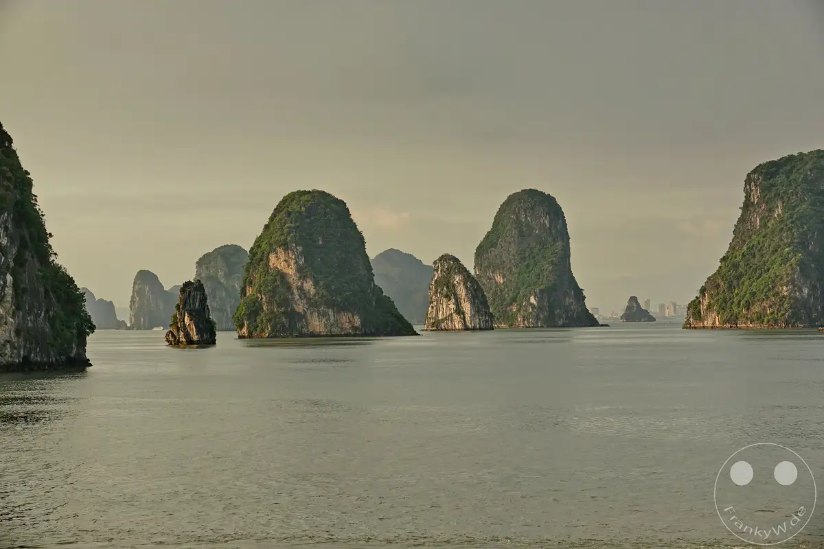 Vietnam und seine berühmte Ha Long Bucht präsentieren sich in einem Postkartenmotiv. Zahlreiche grüne, bewachsene Karstfelsen ragen majestätisch aus dem ruhigen, grauen Wasser empor und bilden eine atemberaubende Insellandschaft. Die Szenerie wird von einem sanften, bewölkten Himmel überstrahlt und vermittelt eine friedliche und geheimnisvolle Atmosphäre.