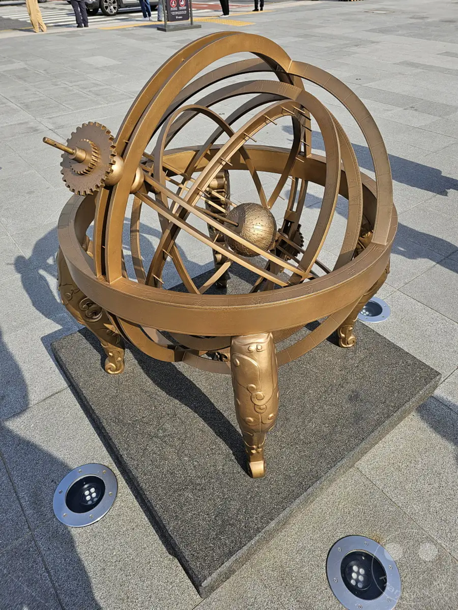 In Seoul, Südkorea, steht auf dem Gwanghwamun Square eine beeindruckende Armillary Sphere, die astronomische Berechnungen veranschaulicht. Das kunstvoll gestaltete Messinstrument aus Bronze ruht auf einem Sockel und ist von einem gepflasterten Platz umgeben. Die Sonneneinstrahlung wirft klare Schatten und betont die geometrische Komplexität der Skulptur, die eine Atmosphäre von Weisheit und Entdeckung vermittelt.