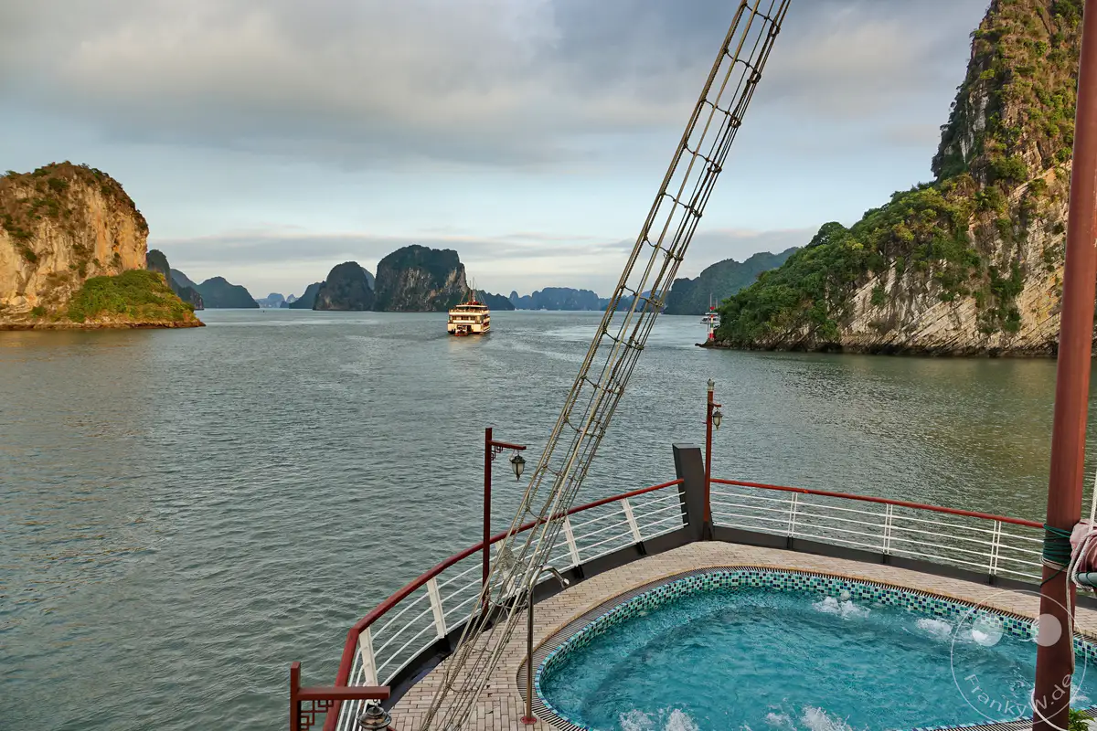 Eine traditionelle Dschunke kreuzt gemächlich durch die malerische Ha Long Bucht in Vietnam, umgeben von majestätischen Karstfelsen, die aus dem türkisfarbenen Wasser ragen. Die untergehende Sonne taucht die Szenerie in ein warmes, goldenes Licht, während eine leichte Bewölkung den Himmel bedeckt und eine ruhige Atmosphäre schafft. Auf dem Deck des Schiffes lädt ein kleiner Pool zur Entspannung ein, während die beeindruckende Naturkulisse die Sinne verzaubert.