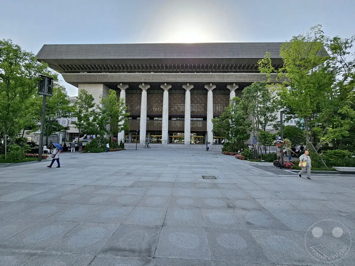 Ein weiter Vorplatz mit großflächigen Steinplatten erstreckt sich vor dem imposanten Sejong Center for the Performing Arts in Seoul, Südkorea. Grüne Bäume säumen die Szene und schaffen einen angenehmen Kontrast zur modernen Architektur des Gebäudes. Einige Personen sind auf dem Platz unterwegs, was dem urbanen Raum Leben einhaucht.
