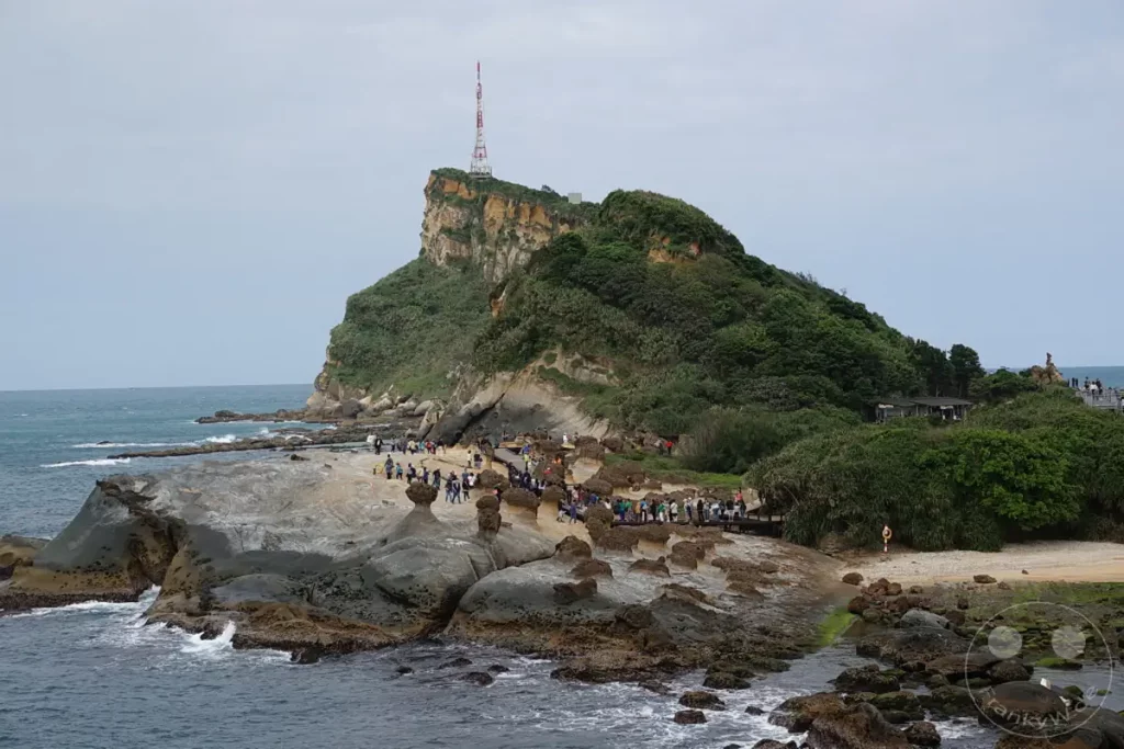 Die Küste Taiwans im Yehliu Geopark bietet eine dramatische Szene mit einzigartigen Felsformationen, die vom Meer umspült werden. Grüne Vegetation bedeckt die steilen Hänge, auf denen sich ein Beobachtungsturm abzeichnet. Touristen erkunden die ungewöhnlichen Gesteinsformationen auf den felsigen Ufern unter einem bewölkten Himmel.