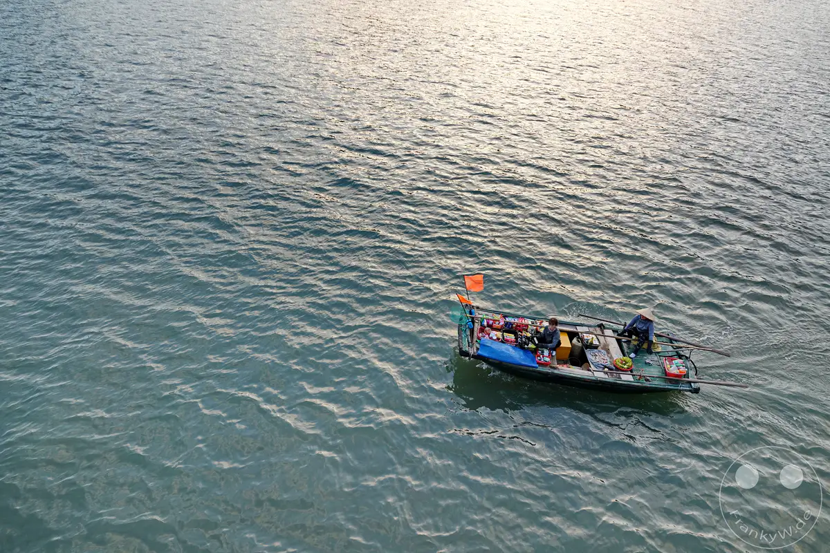 In der Ha Long Bucht Vietnams bietet eine Bootsverkäuferin ihre Waren auf ihrem schmalen Boot an. Das ruhige Wasser reflektiert das sanfte Licht des Himmels und schafft eine friedliche Atmosphäre inmitten des geschäftigen Treiben auf dem Wasser.