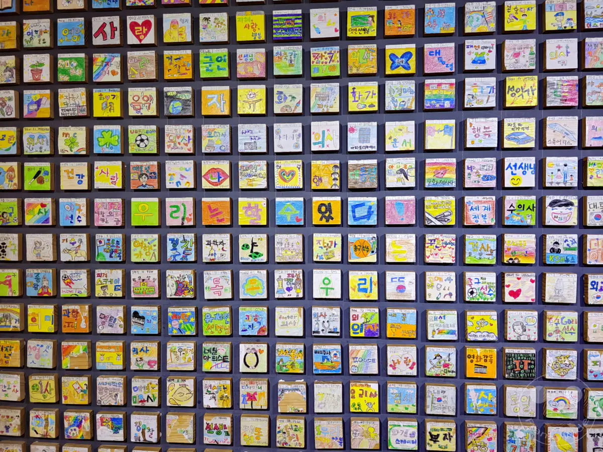 Eine Wand im Sejong Center for the Performing Arts in Seoul, Südkorea, ist mit zahlreichen kleinen, individuell gestalteten Kunstwerken von Kindern bedeckt. Jedes Kunstwerk, oft mit bunten Zeichnungen und handgeschriebenen Texten, schafft ein lebendiges Mosaik kindlicher Kreativität. Die Vielzahl der ausgestellten Stücke strahlt eine Atmosphäre der gemeinschaftlichen Freude und des künstlerischen Ausdrucks aus, die Besucher aller Altersgruppen anspricht.